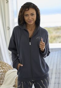 Donkergrijze rits-hoodie met capuchon, voorzakken en geborduurd logo. Zachte stof met een ontspannen pasvorm, gecombineerd met gemusterde legging.