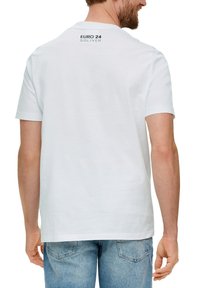 Camiseta de algodón blanca con cuello redondo y mangas cortas. La parte trasera muestra "EURO 24" y "S.OLIVER" en print negro. Jeans de mezclilla visibles.