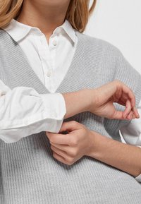 Personne portant une chemise blanche à boutons avec un pull sans manches en tricot gris clair, ajustant le poignet de sa manche de chemise.