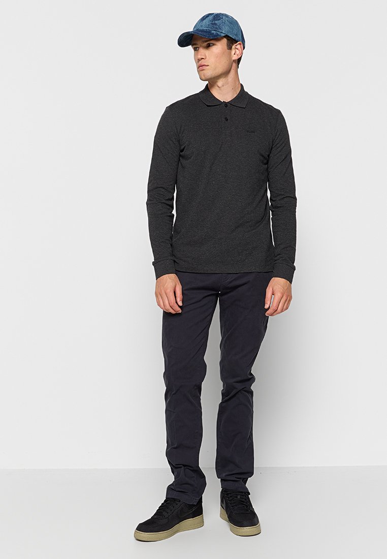 Polo shirt à manches longues gris foncé, col boutonné, associé à un pantalon sombre et des baskets noires. Les accessoires comprennent une casquette en denim bleu.