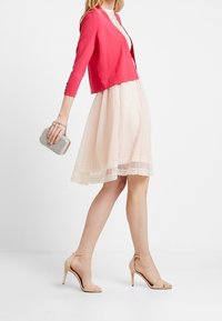 Rosa kofta över en krämfärgad klänning med spetskant, kombinerad med nakna högklackade sandaler och en metallisk kuvertväska. Ren, modern look.