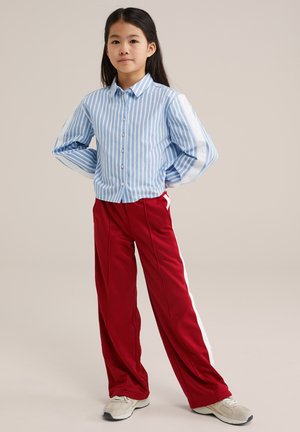 Niña joven con pelo largo que lleva una camiseta a rayas azul y blanca, pantalones deportivos rojos con rayas blancas en los lados y zapatillas blancas, de pie con confianza.