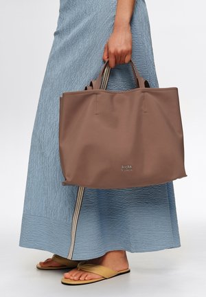 Mano sosteniendo una bolsa de tela marrón con asas cortas, lleva una falda azul texturizada y sandalias planas beige sobre un fondo blanco.