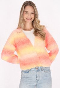 Stickad kofta i gradientnyanser av rosa och gult, med V-hals, löst passform och långa ärmar, i kombination med ljusblå jeans.