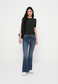 Zwart korte mouwen top met pofschouders, gecombineerd met wijde blauwe spijkerbroek. De outfit wordt aangevuld met witte sneakers en een zwart verenaccent.