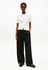 Biały strukturalny kardigan z krótkim rękawem, okrągłym dekoltem i zapięciem na guziki, w połączeniu z czarnymi spodniami o wysokim stanie i szerokich nogawkach oraz błyszczącymi butami.