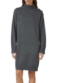 Frau in einem langärmligen, gerippten, dunkelgrauen Strickkleid mit hohem Kragen und Knopfdetails an einer Schulter.