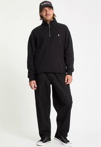 Sort pullover med kvart lynlås, blødt stof, enkelt design med en lille logo. Kombineret med løse, mørke bukser og sorte sneakers.