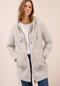 Manteau en polaire gris clair avec fermeture éclair sur le devant, capuche et deux grandes poches avant. Il possède des cordons de serrage et une surface douce et texturée.
