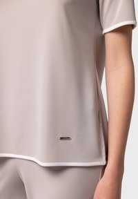 Světlý béžový sportovní top vyrobený z hladkého materiálu. Obsahuje kontrastní bílý lem a malé kovové logo blízko lemu.