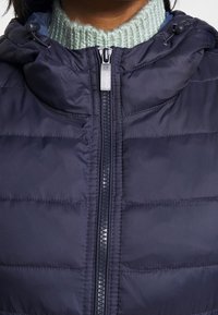 Navyblaue Steppjacke mit horizontalem Quiltmuster und einem Reißverschluss. Verfügt über eine Kapuze mit Kordelzügen und einen hellgrünen gerippten Kragen.
