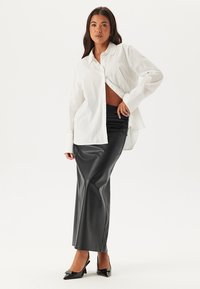 Weißes Button-Up-Hemd mit lockerer Passform, kombiniert mit einem taillierten Rock aus schwarzem Kunstleder und schwarzen spitzen High Heels. Sanfte Texturen überall.