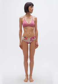 Γυναίκα με κοντά σκούρα μαλλιά, φορώντας ροζ μπικίνι τοπ και μπικίνι με σχέδια λουλουδιών και ψηλή μέση, στέκεται ξυπόλητη μπροστά σε λευκό, απλό φόντο.