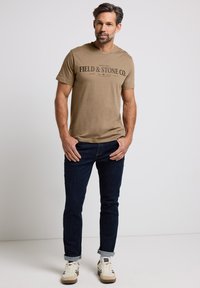 Khaki bavlněné tričko s černým potiskem "FIELD & STONE CO.", spárované s tmavými džínami a bílými teniskami, na neutrálním pozadí.