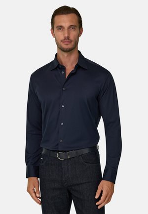 Homme portant une chemise à boutons bleue foncée rentrée dans un jean noir avec une ceinture noire, debout devant un fond uni.