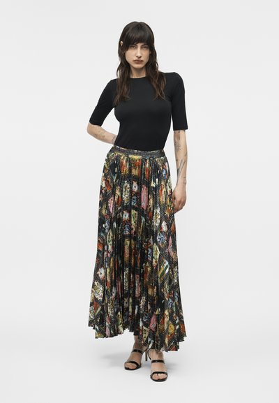 Kobieta w czarnej bluzce z krótkim rękawem i kolorowej, wzorzystej plisowanej długiej spódnicy, ubrana w czarne sandały na obcasie, stoi z jedną ręką na biodrze.