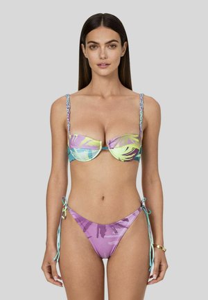 Donna con capelli lunghi e scuri che indossa un bikini tropicale multicolore con braccialetto d'oro al polso sinistro, in piedi davanti a uno sfondo grigio uniforme.