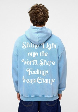 Lichtblauwe hoodie met een voorzijde kangoeroezak, met op de achterkant witte tekst: "Laat Licht op de Wereld Schijnen. Deel Gevoelens. Creëer Verandering."