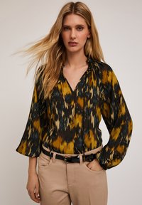 Maglietta a maniche lunghe e scollatura a v con un motivo astratto giallo, nero e blu. Il tessuto cade leggero; abbinata a pantaloni beige e una cintura nera.