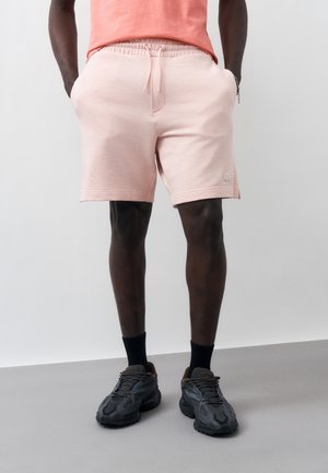 Man wearing light pink drawstring shorts, coral T-shirt, black socks, and gray sneakers standing against a plain background.