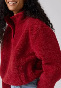 Pulôver de lã vermelha com gola alta, superfície texturada e detalhe de fecho de mola. Apresenta um ajuste descontraído e um acabamento de contraste subtil.