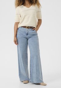 Žena v krémovém pleteném topu s krátkým rukávem, v světle modrých kalhotách s vysokým pasem a širokými nohavicemi, s hnědým opaskem a béžovými botami, stojící s jednou rukou v kapse.