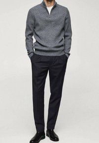 Pull-over gris en maille avec col montant et fermeture éclair au quart, associé à un pantalon tailleur foncé et des bottines noires.