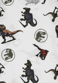 Biała koszula z różnorodnymi nadrukami dinozaurów w różnych kolorach, w tym zielonym, pomarańczowym i czarnym, z okrągłymi akcentami logo Jurassic.