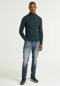 Tamnozelena pletena turtleneck majica, uparena s izlizanim svijetloplavim farmerkama i sivim tenisicama, s malim logotipom na prsima.