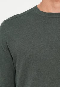 Mörkgrön tröja med ribbad textur, rund halsringning och insydda ärmar. Tyget verkar mjukt och lätt.