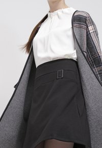 Valkoinen paita, jossa on ryhmitelty kaula-aukko, harmaa ruudullinen takki ja mustat korkeavyötäröiset hameet, joissa on solkilukko-tietoa. Suuret tekstuurit ja kerroksellinen muotoilu.