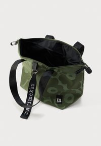 Zaļš auduma iepirkumu maisiņš ar ziedu rakstu, melnu rāvējslēdzēju, melnām rokturiem un melnu siksnu ar uzrakstu "marimekko", atvērts, lai parādītu melnu iekšējo oderi.