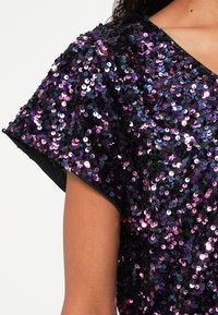 Sort stoftop dækket af lilla og sorte palietter, med en bred halsudskæring og korte ærmer, der fremhæver en reflekterende tekstur.