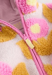 Veste zippée en polaire avec des motifs floraux surdimensionnés roses et jaunes sur un fond crème, dotée d'une fermeture éclair rose et d'un tissu texturé.