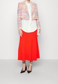 Giacca multicromatica tagliata con trama testurizzata sopra una camicia bianca, abbinata a una gonna a coste rossa brillante e tacchi a punta neri.