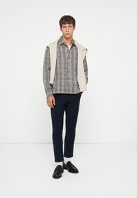 Grau kariertes Hemd mit Knöpfen, darüber ein beiger Sweater-Weste, navy Slim-Fit-Hose und schwarze Slipper, stehend vor einem weißen Hintergrund.