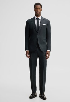 Homme debout, droit, portant un costume sombre sur mesure avec une chemise blanche, une cravate noire, des chaussures habillées noires et un mouchoir blanc dans la poche.