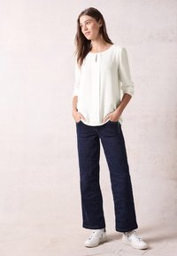Witte top met lange mouwen en een sleutelgatdetail, gecombineerd met marineblauwe wijde broek en witte sneakers, op een lichte neutrale achtergrond.