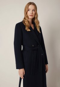 Granatowy wełniany płaszcz o podwójnym zestawieniu, z przednimi kieszeniami i materiałowym paskiem. Posiada klapowy kołnierz oraz gładką teksturę.