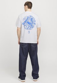 Camiseta blanca de algodón con un diseño gráfico azul de un pájaro rodeado de intrincados remolinos, combinada con pantalones de mezclilla oscura.