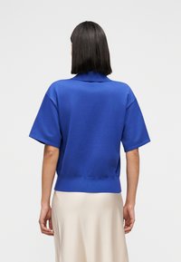 Ρουά μπλε πόλο μπλουζάκι με κοντό μανίκι και ριμπ τελείωμα, με γιακά και λείο ύφασμα. Φορεμένο πάνω από φούστα ανοιχτού χρώματος.