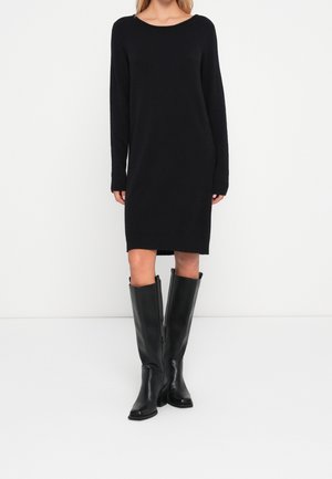 Femme portant une robe en maille noire à manches longues associée à des bottes en cuir noires montantes sur un fond blanc uni.