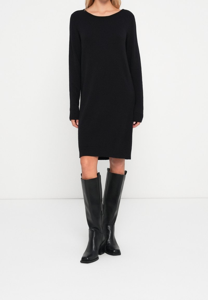 Femme portant une robe en maille noire à manches longues associée à des bottes en cuir noires montantes sur un fond blanc uni.