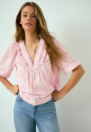 Žena s dlhými svetlohnedými vlasmi má na sebe ružovú blúzku s čipkovými detailmi a modré džínsy, stojí s rukami v bok proti svetlému pozadiu.