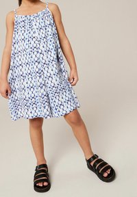 Dziecko ubrany w bezrękawnikową sukienkę w niebiesko-białe wzory, z czarnymi sandałami na paski, stojące z jedną nogą nieco wysuniętą do przodu.