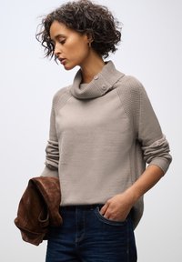 Ljusbeige tröja med en cowl-krage och knapppdetaljer. Texturerade accenter på axlarna. Modellen håller en brun mockajacka. Avslappnad stil.