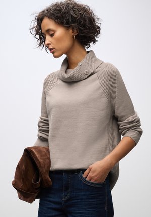 Suéter beige claro con cuello de cisne y detalle de botones. Acentos texturizados en los hombros. La modelo sostiene una chaqueta de ante marrón. Estilo casual.