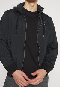 Chaqueta bomber ligera negra con cierre de cremallera, capucha ajustable, puños acanalados y bolsillos. Se lleva sobre una camiseta negra. Tejido texturizado.