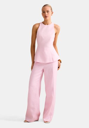 Vrouw draagt een mouwloze lichtroze top en bijpassende wijde broek, staand tegen een witte achtergrond.