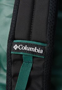 Black mesh backpack strap with green accents, featuring a prominent white "Columbia" logo tag. Textured surface provides grip.